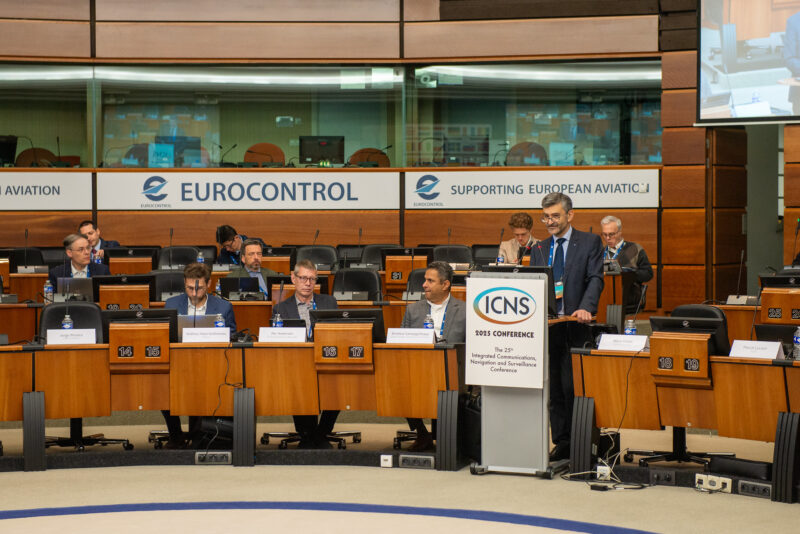 A speaker stands at a podium labeled ICNS in a conference room. Seated attendees are at curved desks. Behind them, a sign reads EUROCONTROL and Supporting European Aviation. Multiple monitors and microphones are visible.